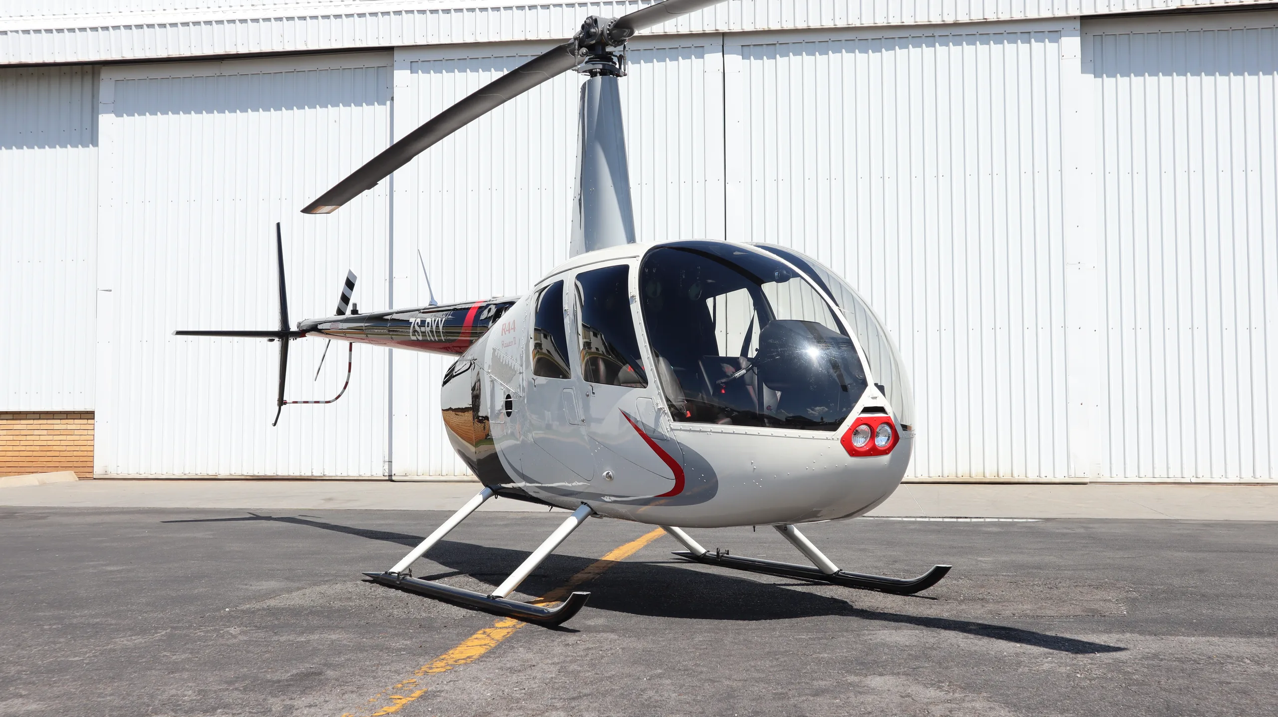 A white and black Robinson R44 Raven II helicopter with red accents is parked on an asphalt tarmac in front of a white hangar.