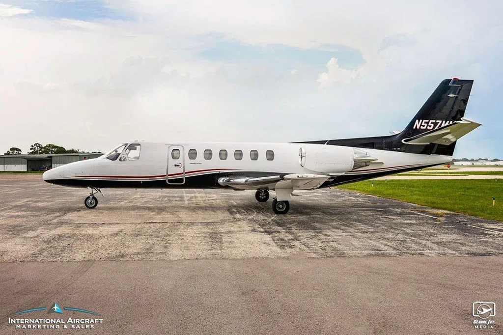 A side view of a white and black Cessna Citation business jet (N557MM) parked on an airfield under a cloudy sky.