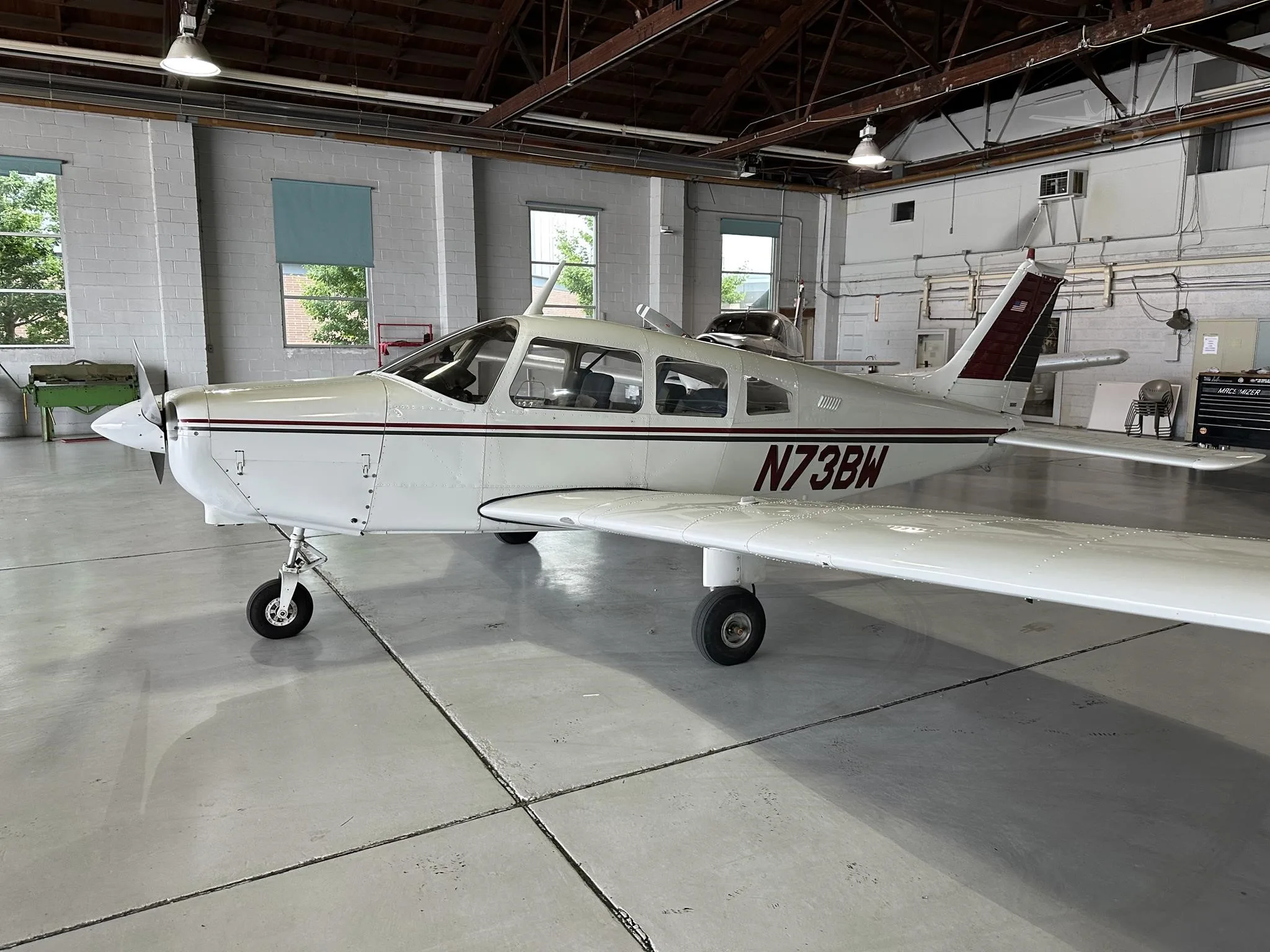 A white single-engine aircraft with maroon and black stripes, registration N73BW, viewed from the front-left in a hangar.