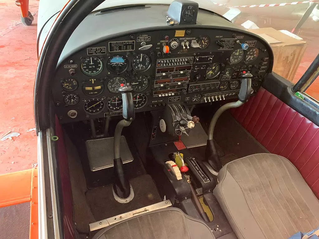 A wide view of the aircraft's cockpit showing the instrument panel, control sticks, rudder pedals, and a grey pilot's seat.