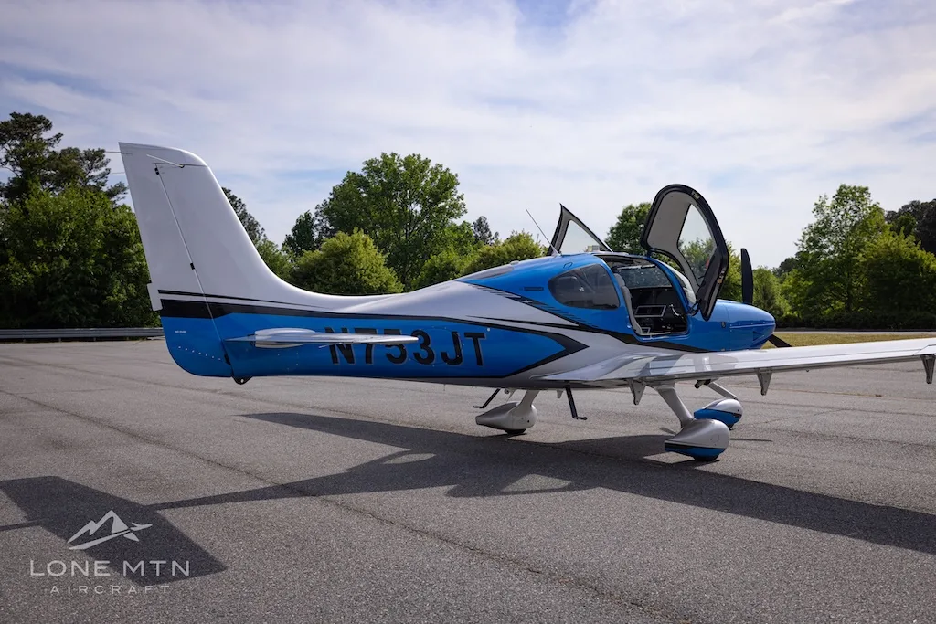 A blue and silver single-engine aircraft, N753JT, with open gull-wing doors is parked on an asphalt tarmac with trees in the background.