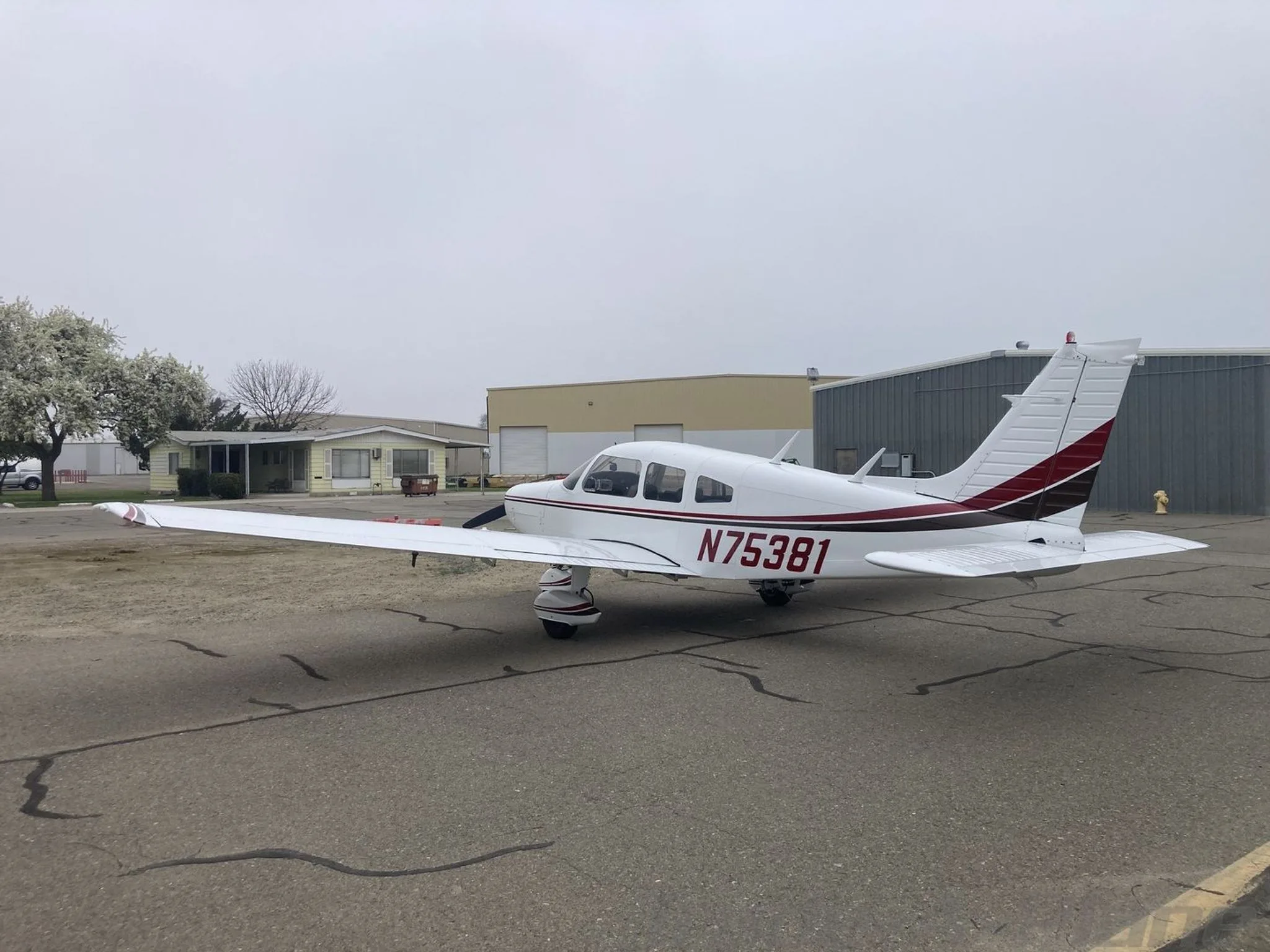 A white Piper PA-28-151 aircraft with red and black stripes parked on an airfield with buildings and trees in the background.