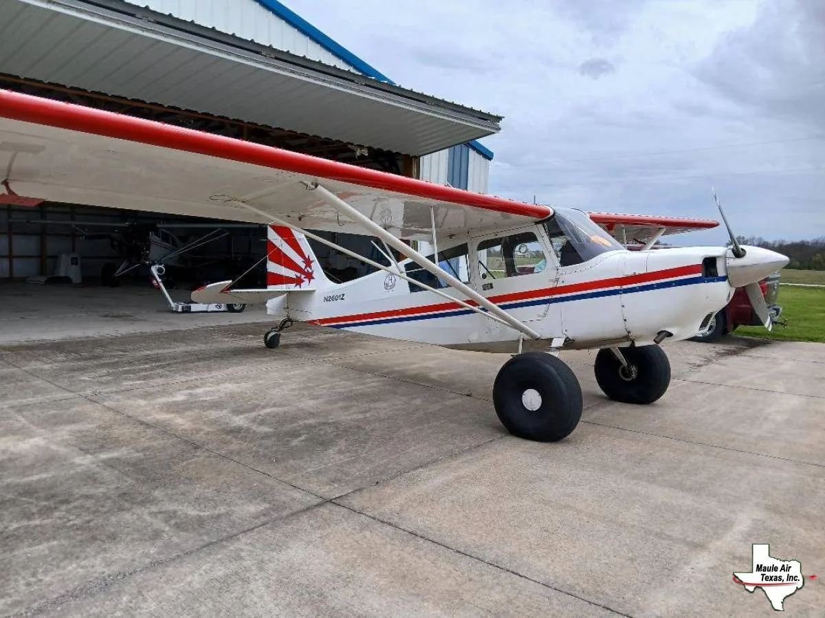 A white high-wing Maule aircraft with red and blue stripes and large bush tires, parked on a concrete apron next to a hangar.