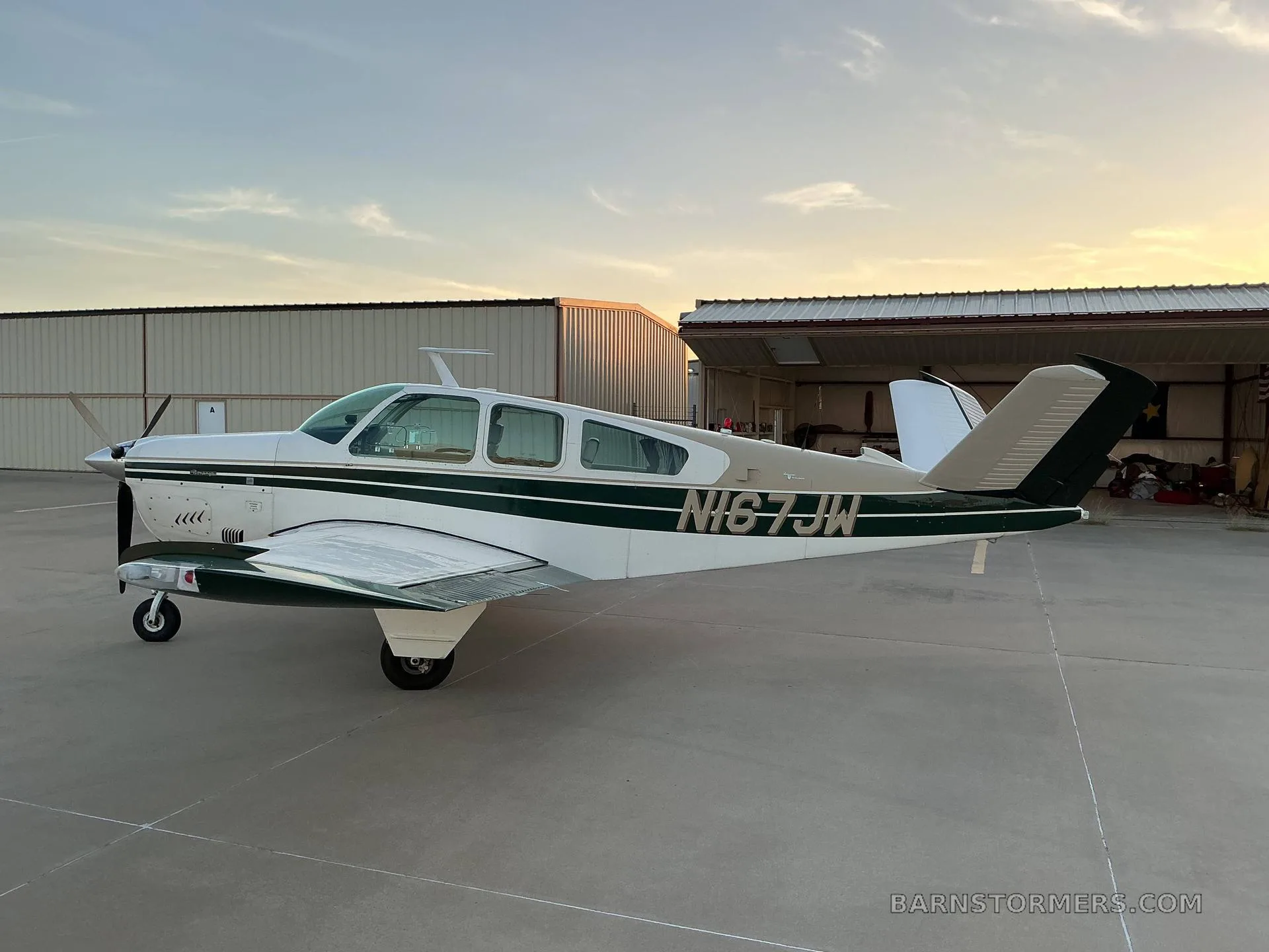 A white and dark green Beechcraft Bonanza (N167JW) parked on an airfield tarmac at sunset, with hangars in the background.