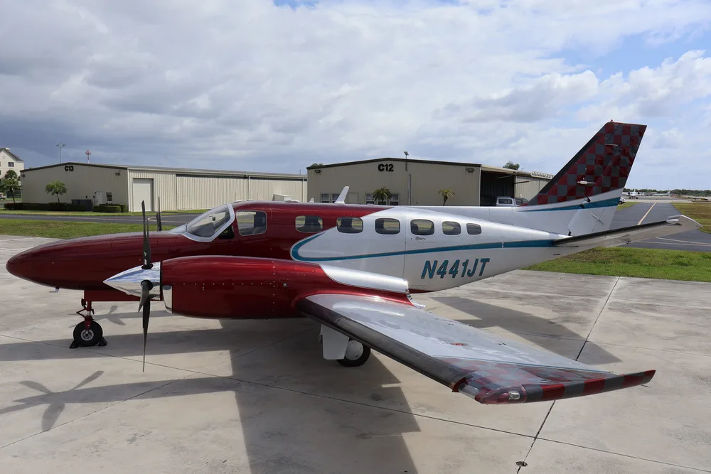 A side view of the red and silver twin-engine aircraft N441JT parked on an airfield next to hangars.
