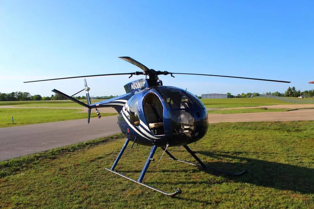A blue and white Hughes 500 helicopter (N500RT) sits on a grassy field next to a runway under a clear blue sky.
