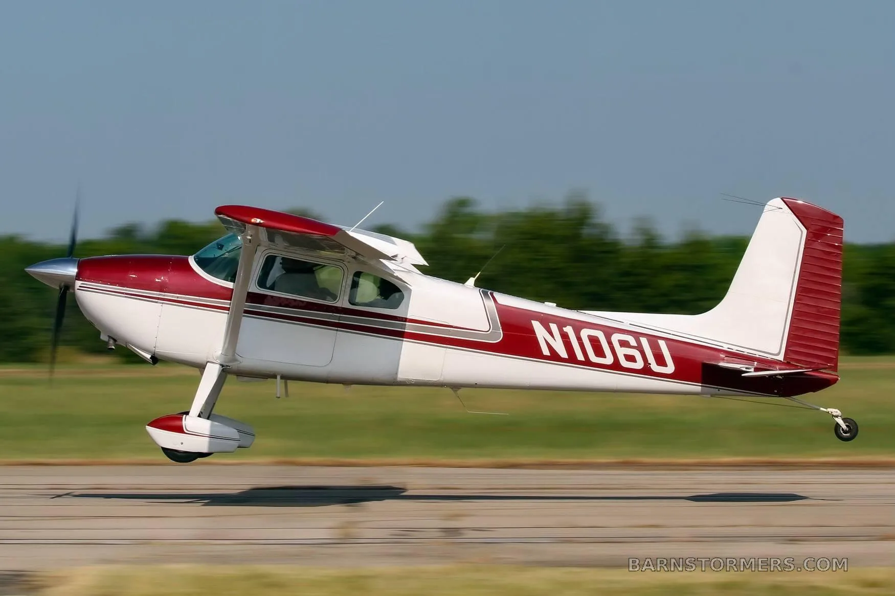 A white and maroon Cessna 182 aircraft (N106U) is captured in flight, low over a runway with a green background.