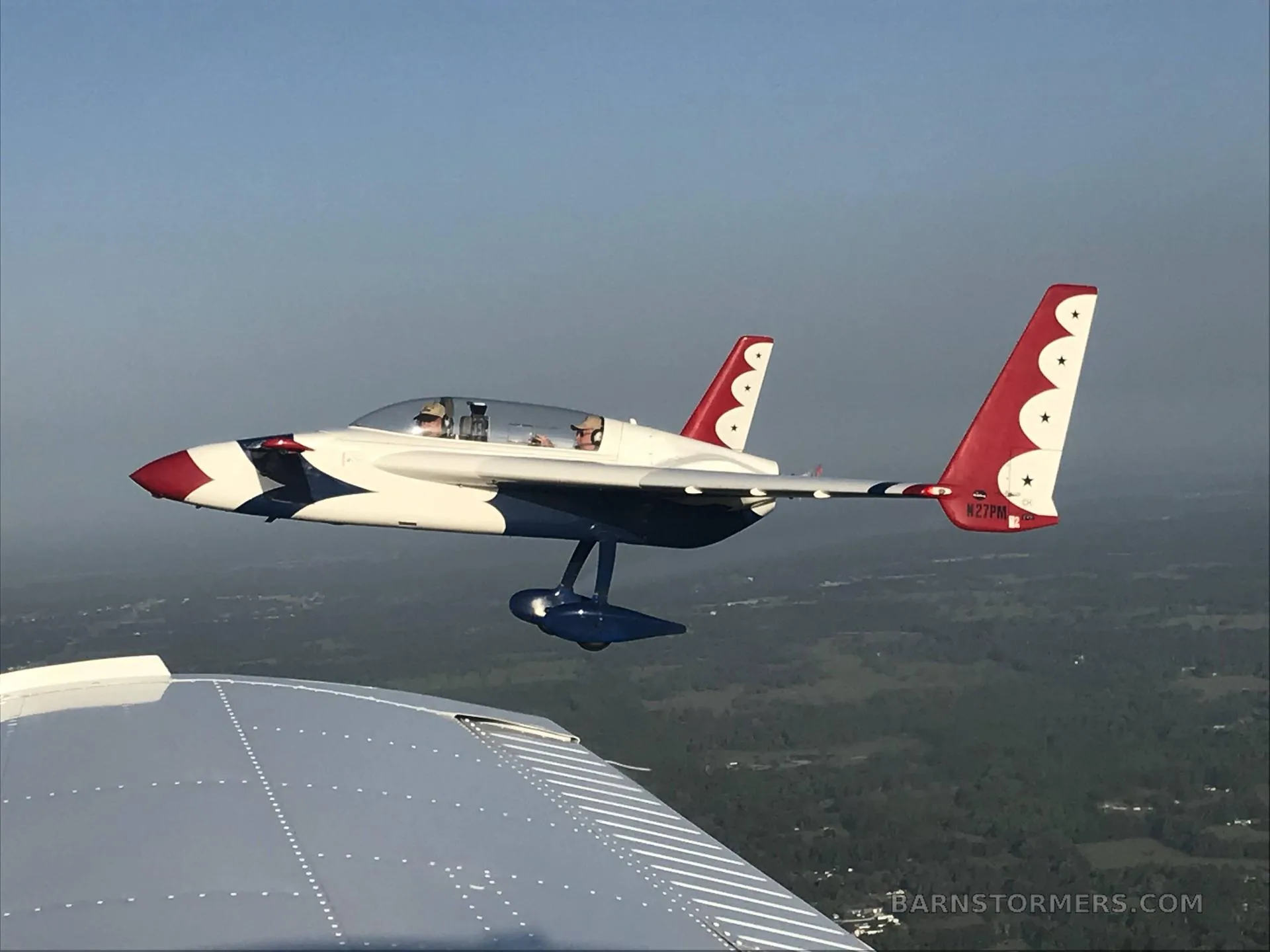 A white, red, and blue aircraft with twin tails and a pusher propeller, seen in flight from another aircraft's wing, with a pilot visible in the cockpit.