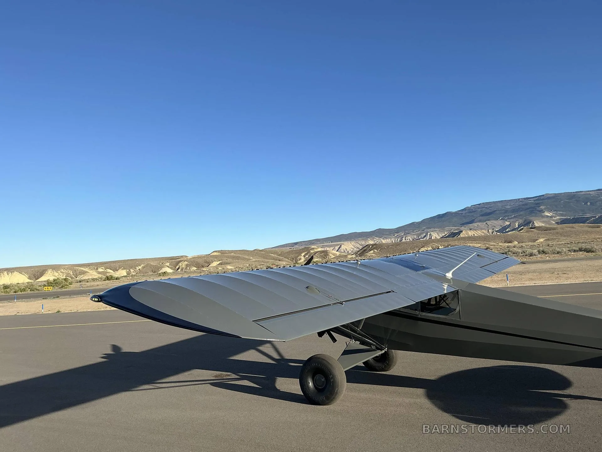 A close-up view of the right wing and tail of a dark grey Carbon Cub FX3 aircraft on an airfield.