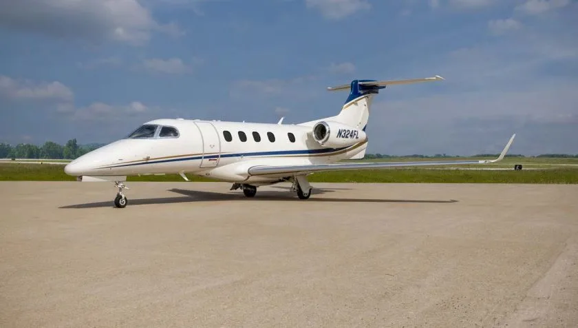 A white Embraer Phenom 300 private jet with blue and gold stripes on the tail and fuselage, parked on an airfield under a partly cloudy sky.
