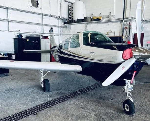 A front-side view of a dark blue and white Beechcraft Bonanza N36HC with red propeller covers in a hangar.
