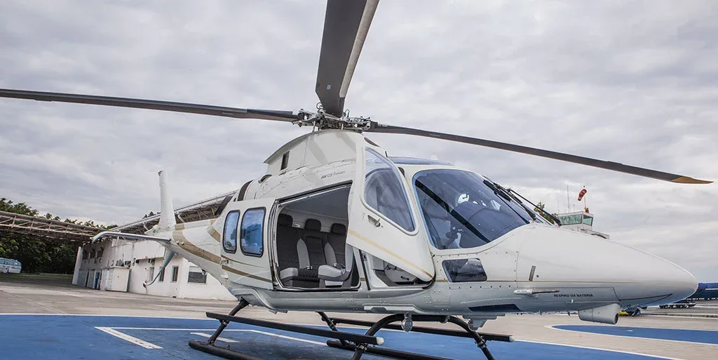 A white helicopter with gold stripes is parked on a blue helipad with its front and cabin doors open, viewed from a front-side angle.
