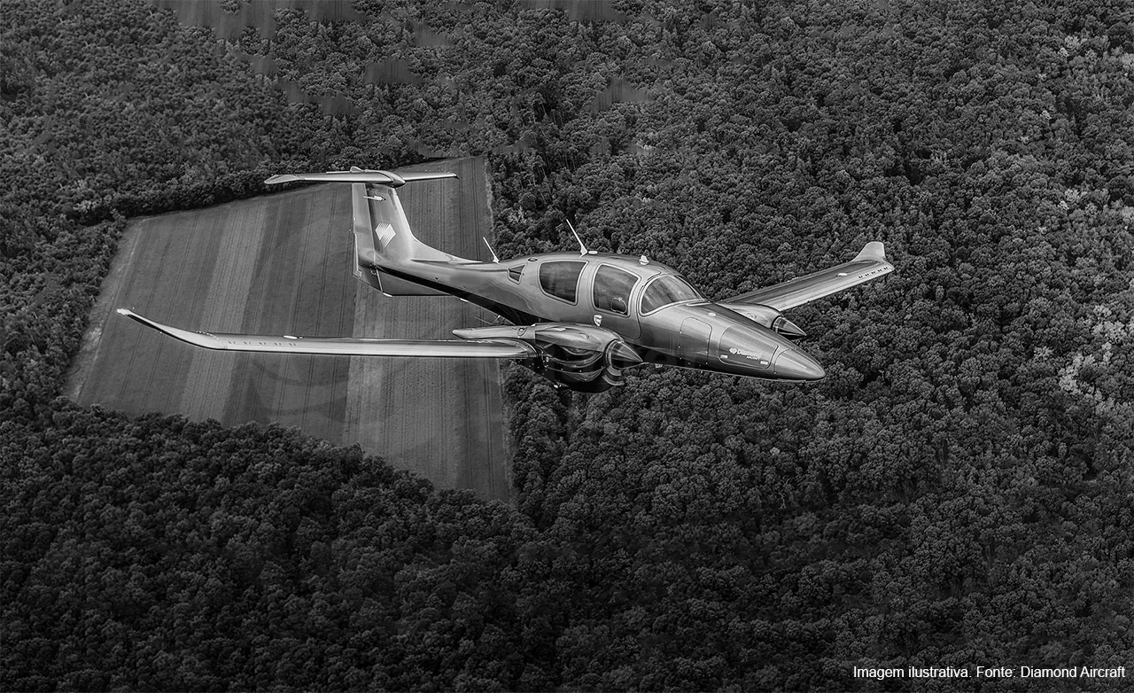 A black and white aerial view of a Diamond DA62 aircraft flying over a forest and field.