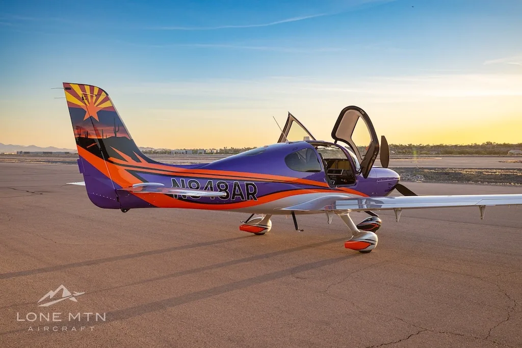 Side view of a purple and silver Cirrus SR22T with open doors, featuring an Arizona flag tail design, parked on a tarmac at sunset.