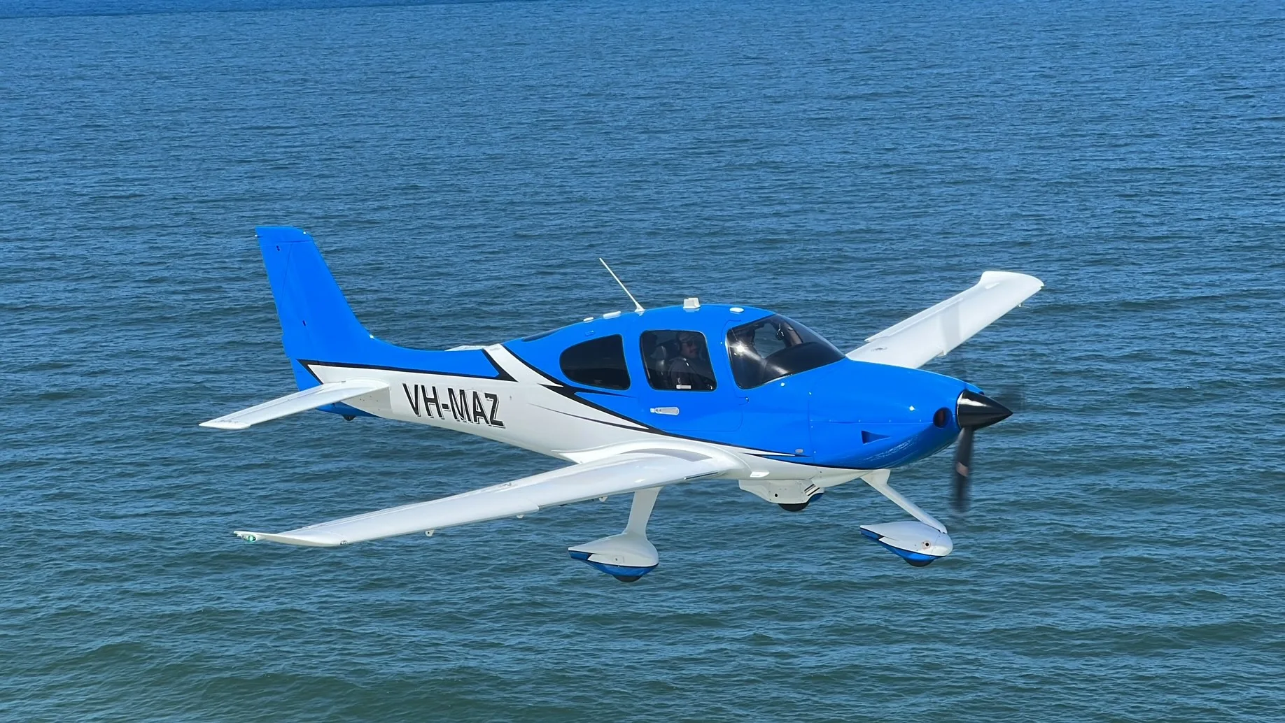 A blue and white single-engine propeller aircraft, registration VH-MAZ, flying low over blue water.