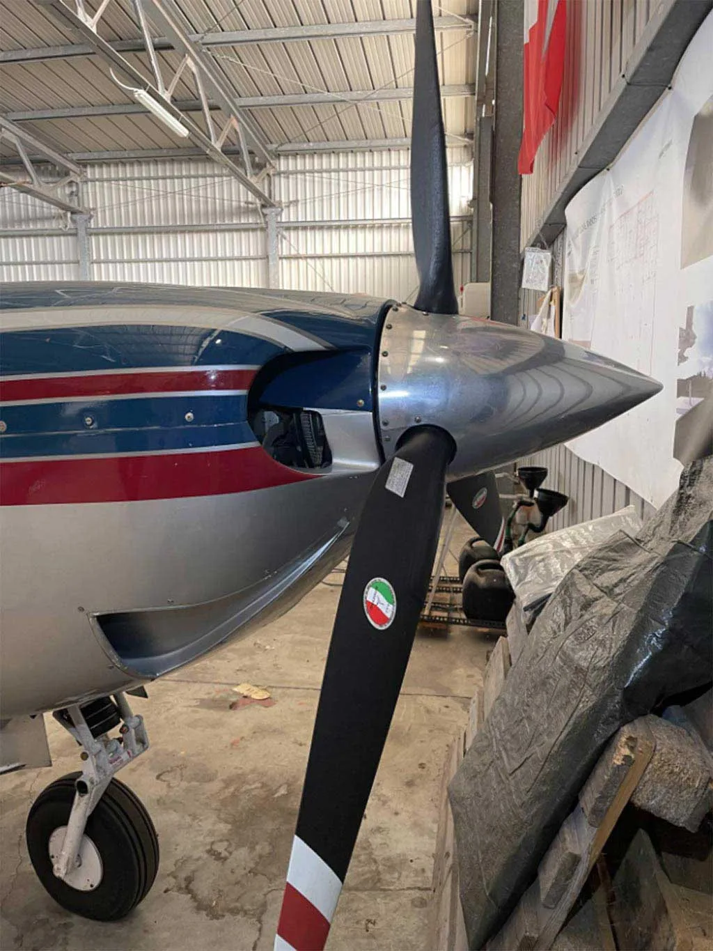 A close-up view of the propeller and nose of a Mooney M20J aircraft, showing its silver, blue, and red paint scheme inside a hangar.