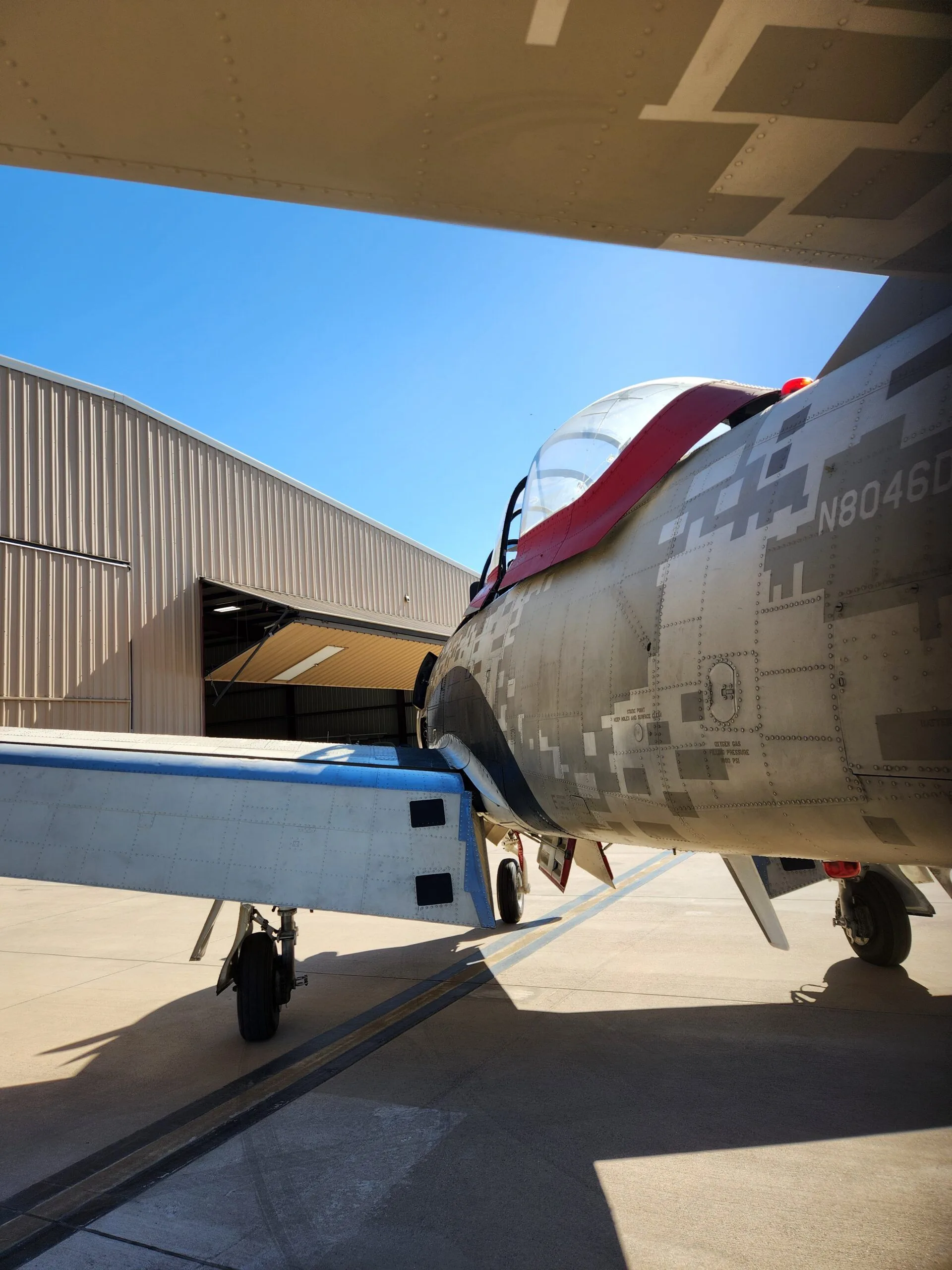 The tail section of a T-28 Trojan aircraft, showing its N-number N8046D and desert digital camouflage paint scheme, parked on an airfield.