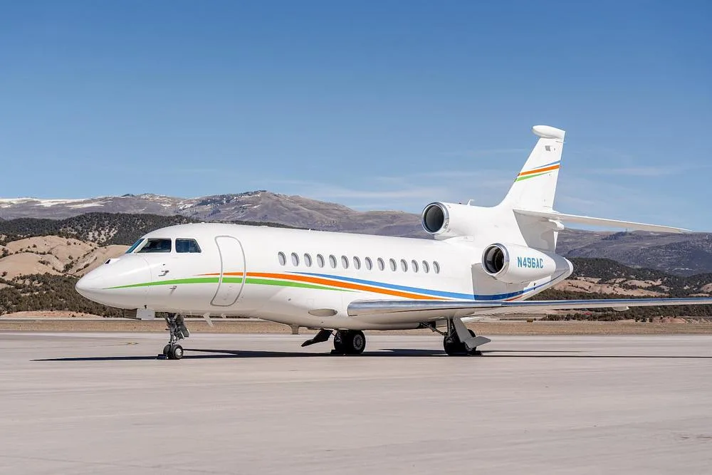 A white Dassault Falcon 900EX, registration N496AC, with green, orange, and blue stripes, parked on an airfield with mountains in the background.