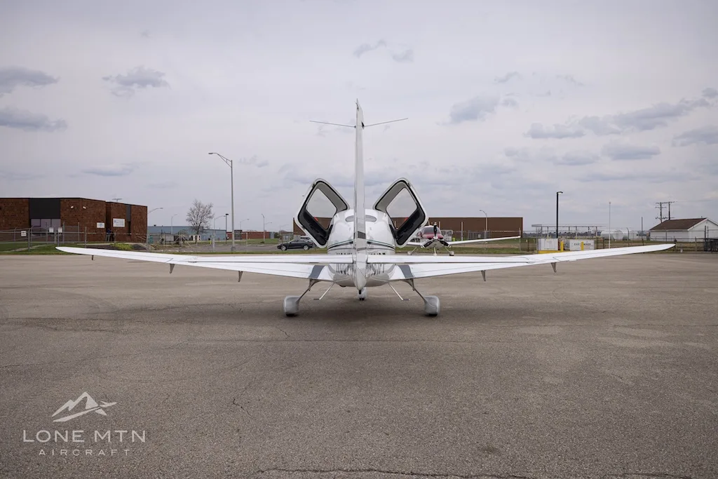 A rear view of a white Cirrus SR22 aircraft with its doors open, parked on an asphalt ramp.