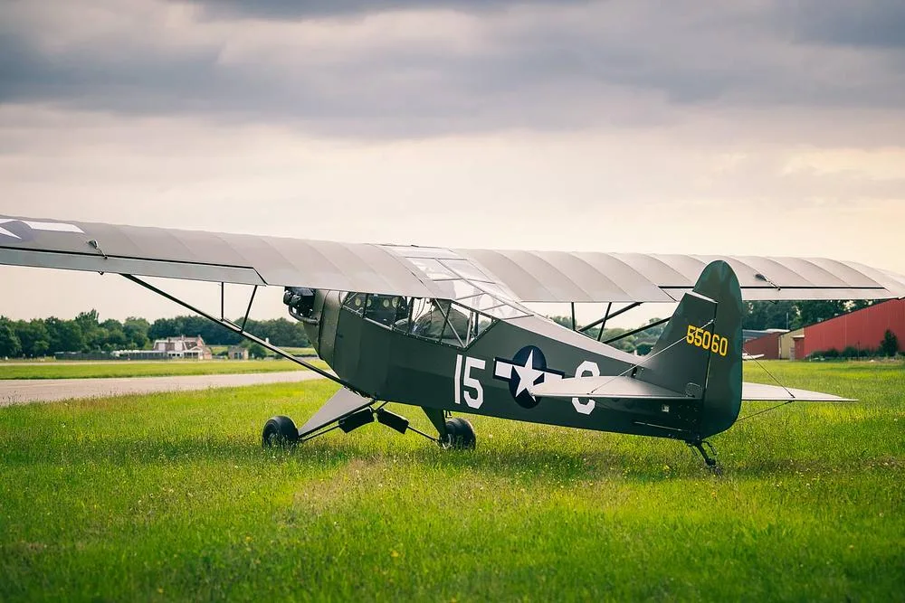 A Piper L-4 Grasshopper in olive drab military livery, with "55060" on the tail, parked on grass next to a paved runway under a cloudy sky.