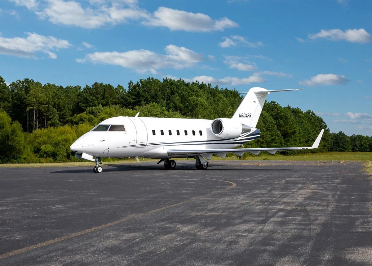 A white Bombardier Challenger 604 business jet (N604PS) with blue stripes on the tail, parked on an asphalt tarmac with trees in the background under a blue sky.
