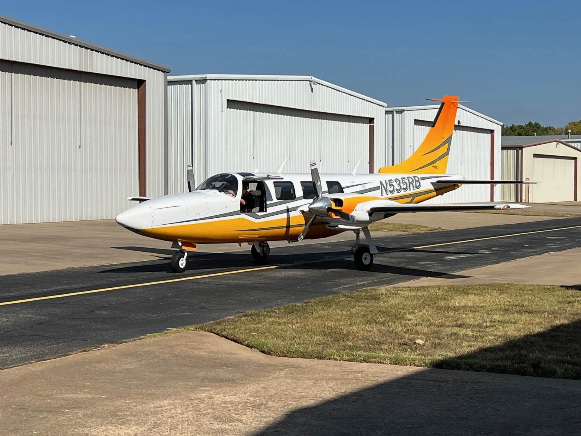 A white and orange twin-engine Piper Navajo aircraft (N535RB) taxiing on an asphalt runway with hangars in the background.