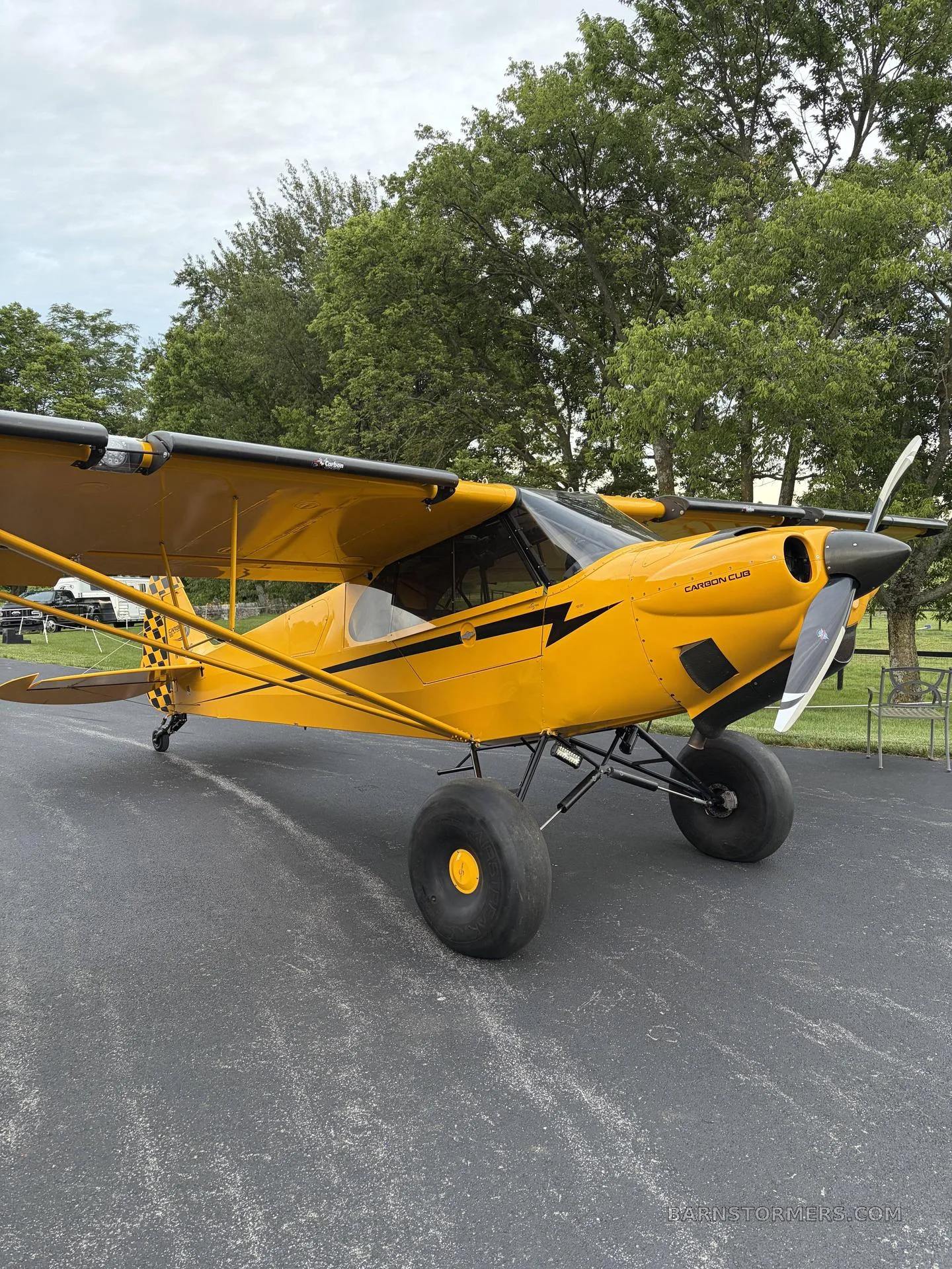 A yellow Carbon Cub aircraft with large tundra tires is parked on an asphalt driveway with trees in the background.