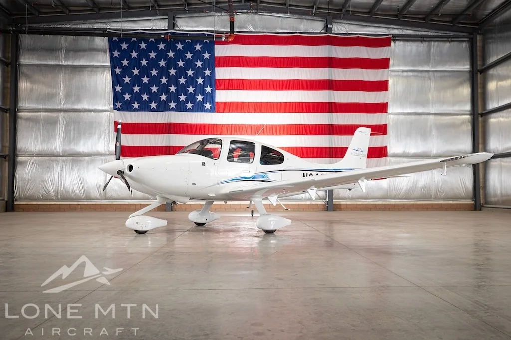 A white Cirrus SR22 aircraft with blue and black stripes, tail number N643LV, parked in a hangar with an American flag in the background.