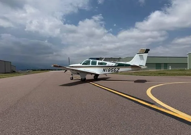 A white and gold Beechcraft Bonanza aircraft, tail number N1856Z, is parked on an airfield next to a yellow taxiway line.