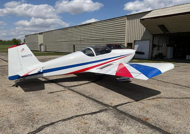 A white single-engine aircraft with red and blue stripes, parked on a tarmac next to an open hangar.