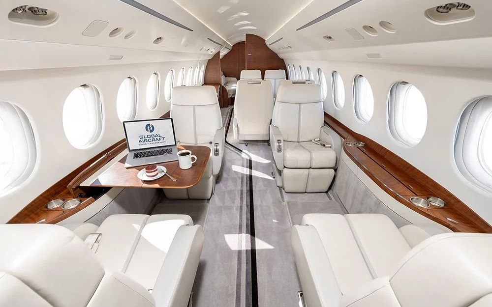 The luxurious interior cabin of a private jet with light-colored leather seats, a wooden table, and a laptop.