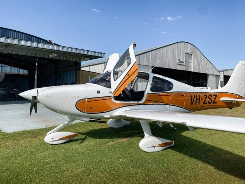 A white and orange single-engine propeller aircraft with gull-wing doors open is parked on grass in front of hangars.