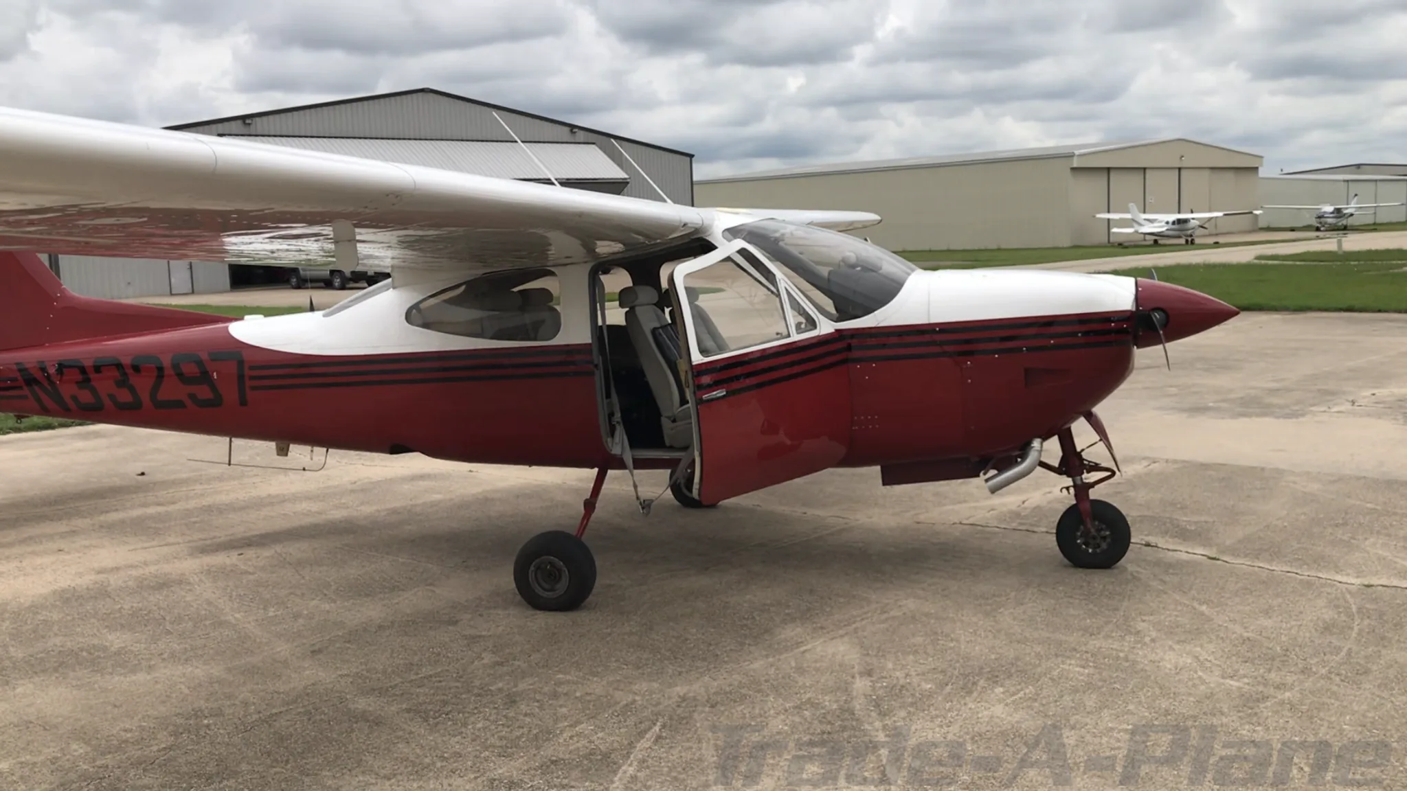A side view of the red and white N33297 aircraft with its door open, parked on an airfield with other aircraft and hangars in the background.