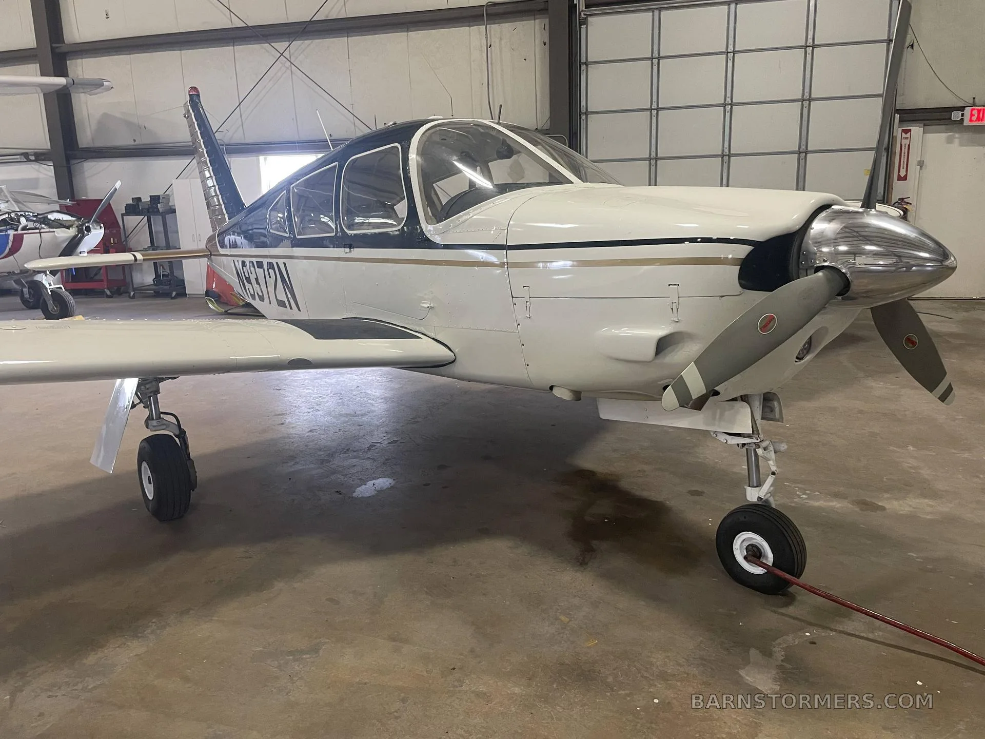 A white and black single-engine aircraft with a gold stripe, registration N9372N, parked in a hangar, viewed from the front-right.