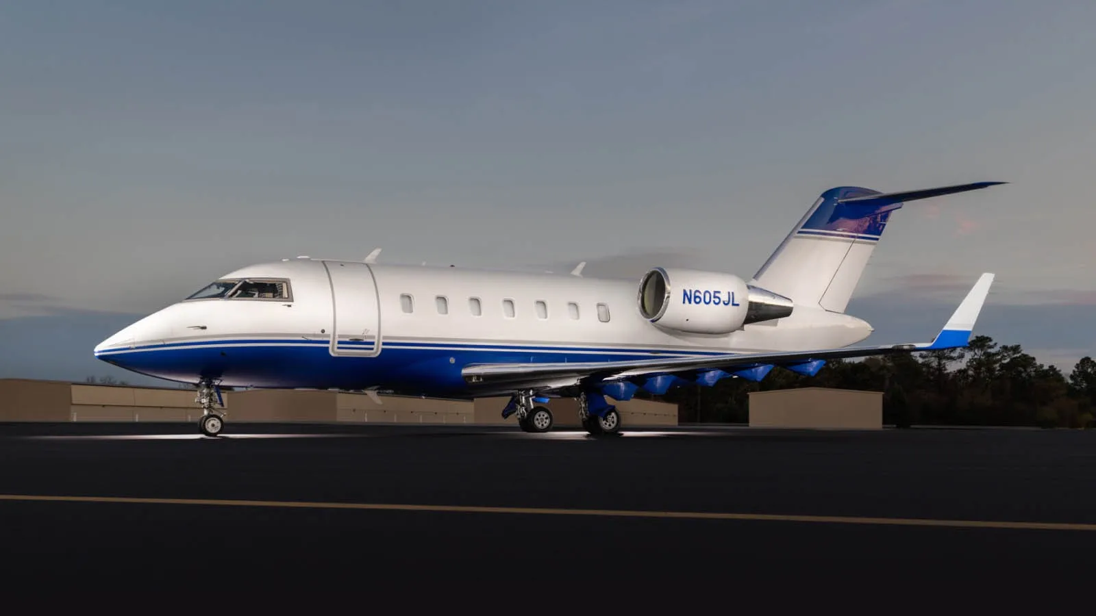 A Bombardier Challenger 605 business jet, registration N605JL, with a white and blue paint scheme, parked on a tarmac at dusk.