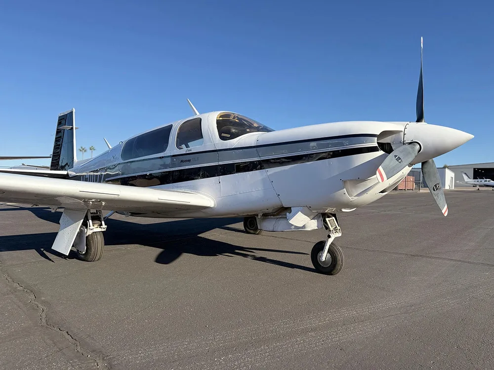 A front-right view of a white Mooney aircraft with black and grey stripes, tail number N711WZ, parked on an airfield.