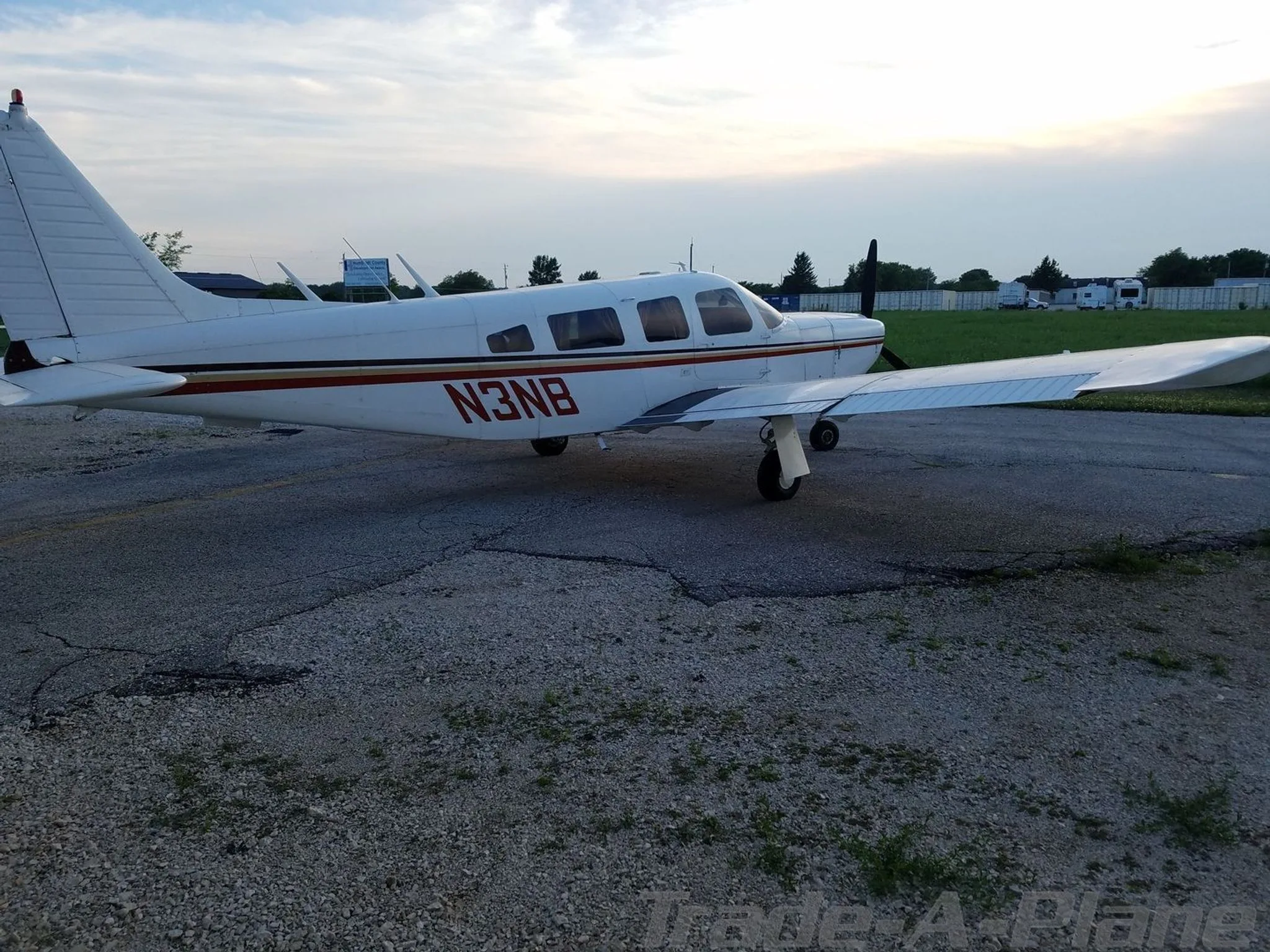 A white Piper aircraft with red and brown stripes, registration N3NB, parked on an airfield at dusk.