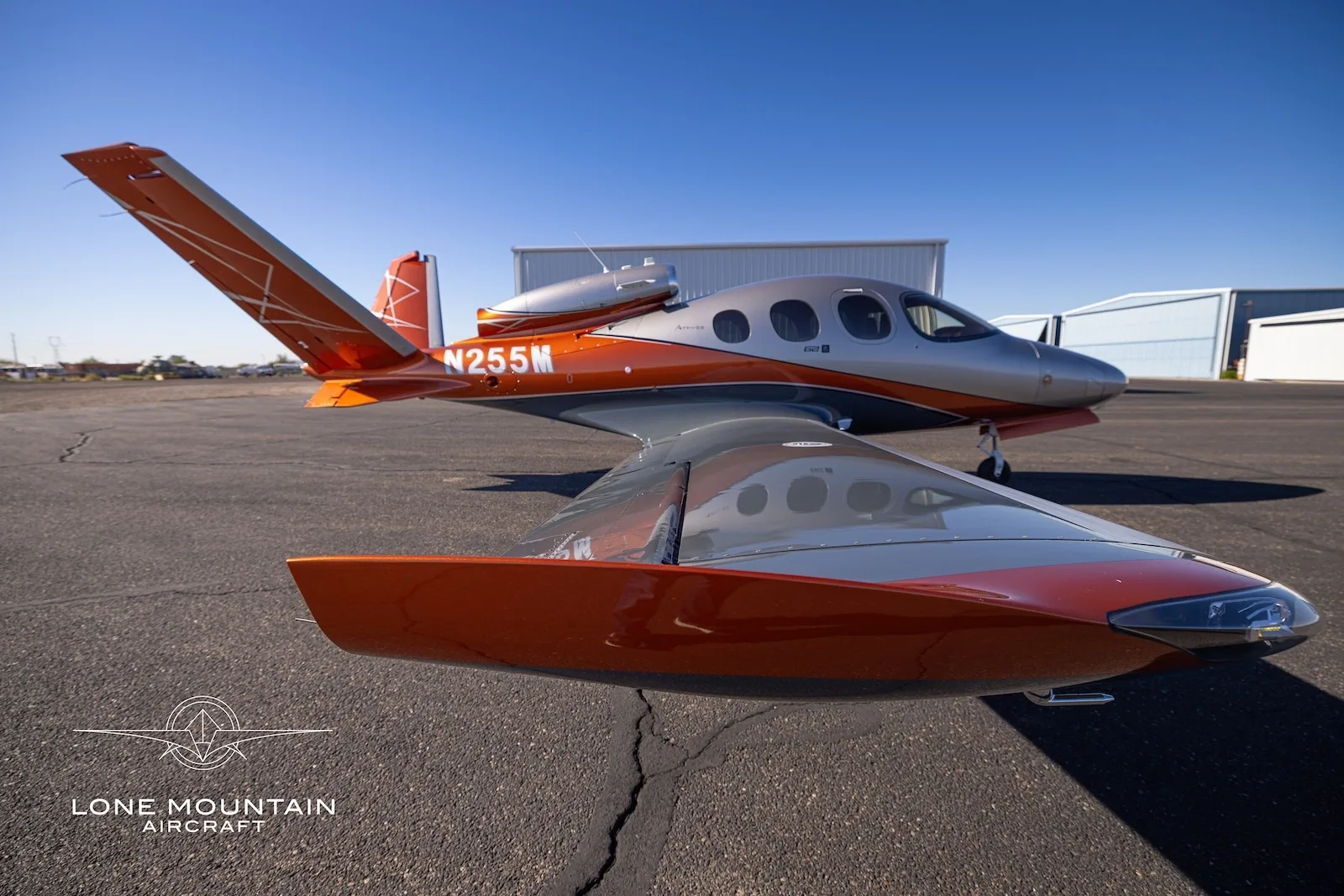 A Cirrus Vision Jet (SF50) with registration N255M, featuring a silver and orange paint scheme, parked on an asphalt tarmac.