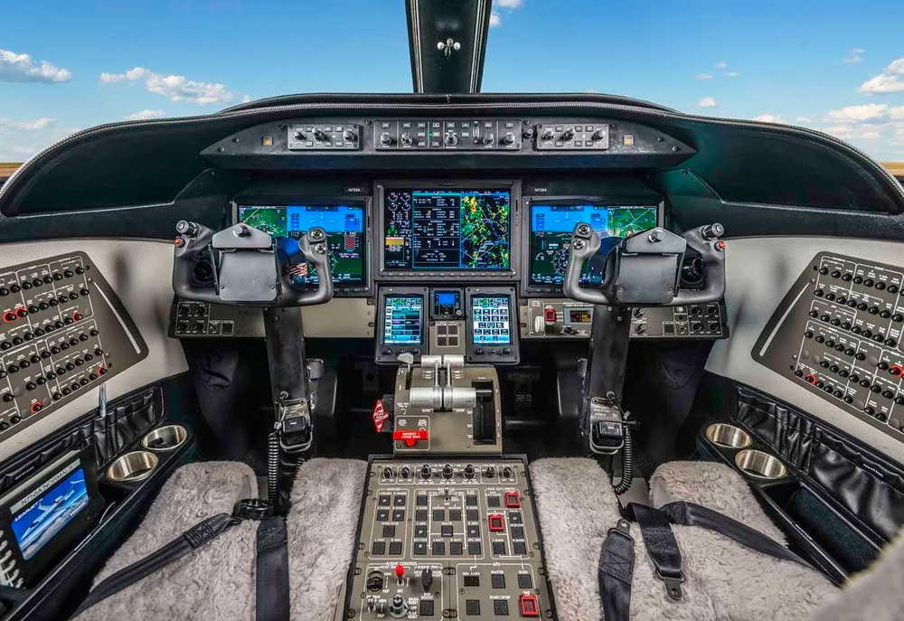The cockpit of a private jet displays multiple digital screens, control yokes, and numerous buttons and switches.