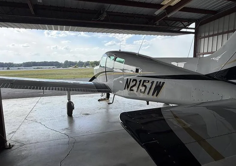 The tail section of a white and black single-engine propeller aircraft, N2151W, is parked inside a hangar with an airfield visible outside.