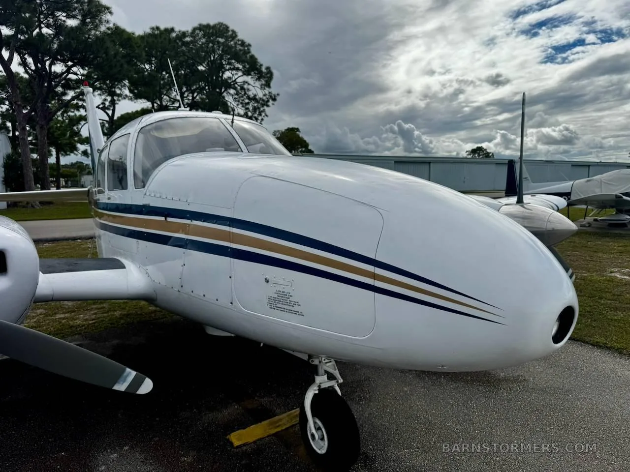 A close-up side view of the nose and front fuselage of a white twin-engine aircraft with blue and gold stripes, showing the landing gear.