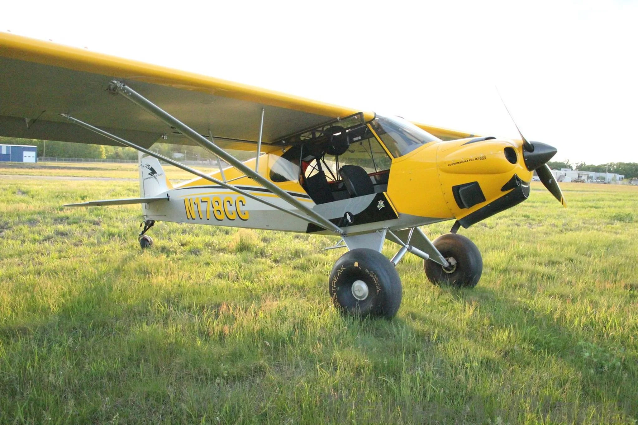 A yellow and grey Carbon Cub aircraft, N178CC, with large tundra tires, parked on a grassy field.