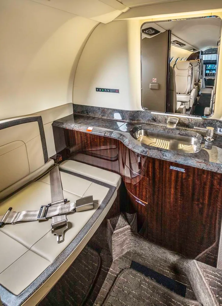 A private jet lavatory features a beige seat with a seatbelt, a granite countertop with a sink, and a mirror.