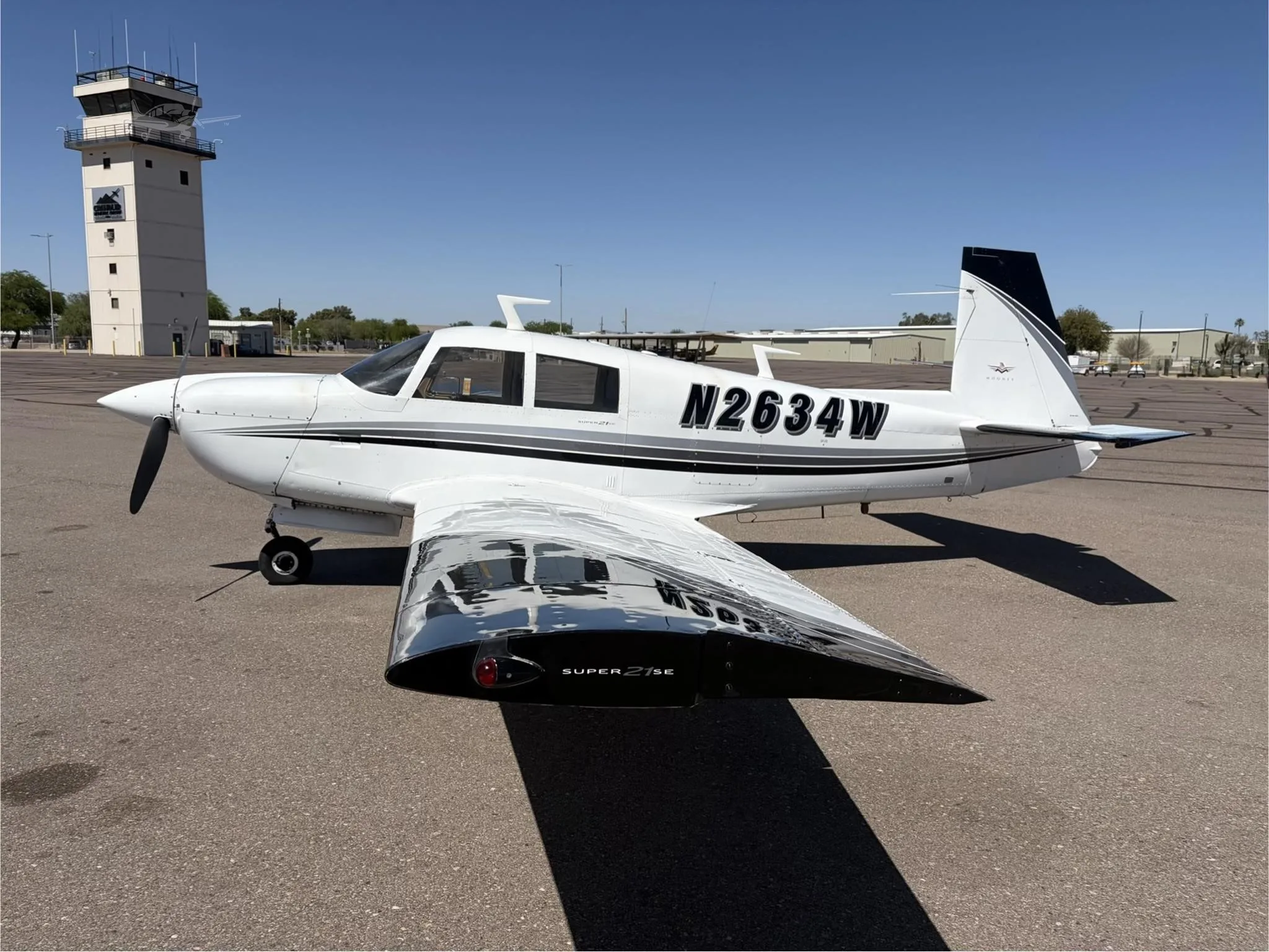 A white single-engine propeller aircraft with black and grey stripes, tail number N2634W, parked on an airport tarmac with a control tower in the background.