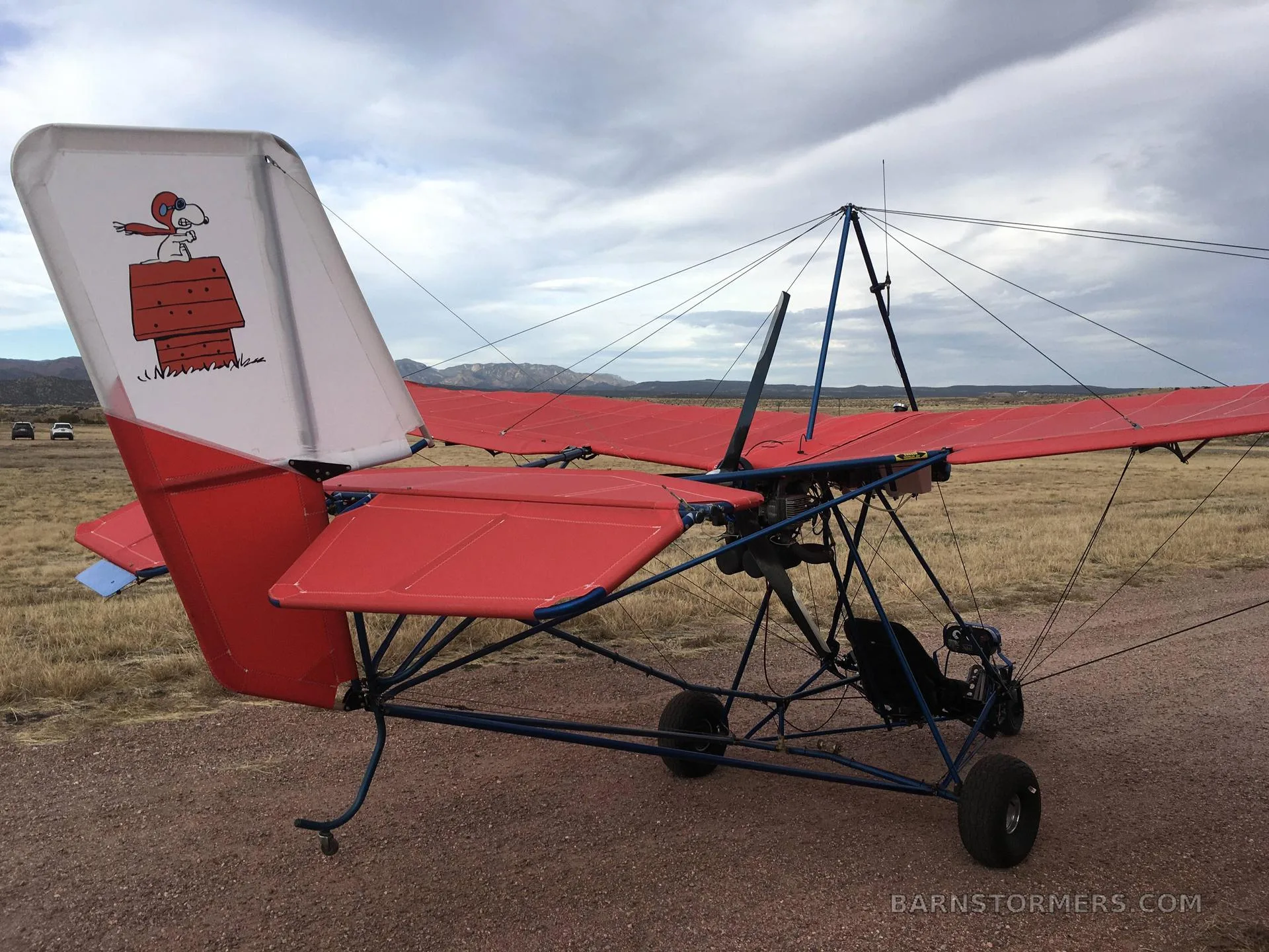 A red ultralight aircraft with a Snoopy graphic on its tail, parked on a dirt path with mountains in the background.