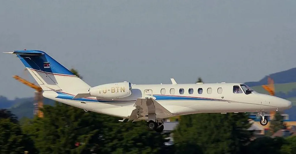 A white business jet with blue and gold stripes, registration YU-BTN, in flight with landing gear extended, against a background of trees and hills.