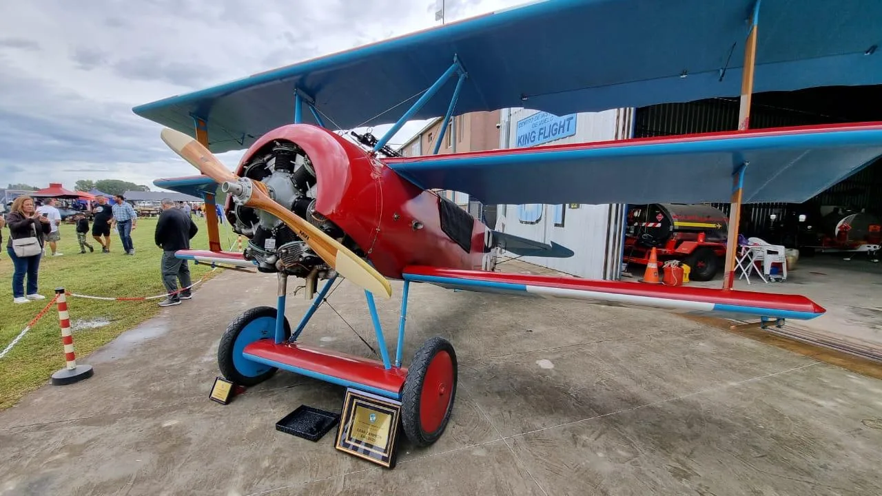 A red and blue Fokker Dr.I replica triplane with its engine exposed, parked on concrete at an outdoor event with people in the background.