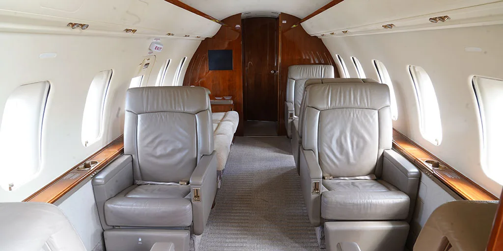 The interior cabin of a private jet features light grey leather seats, a beige carpet, and rich wooden paneling along the walls and a rear bulkhead.