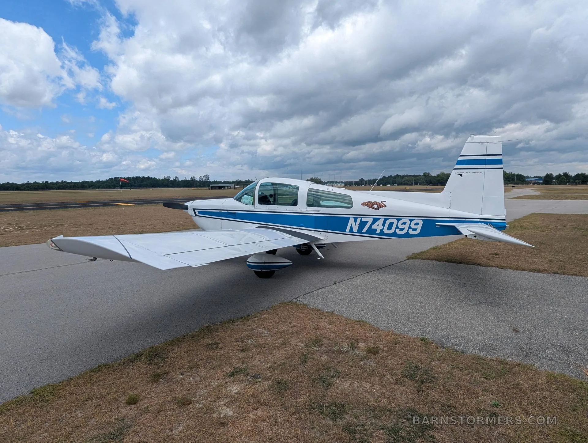 A white and blue Grumman AA-5 aircraft with registration N74099 parked on an asphalt ramp under a cloudy sky.