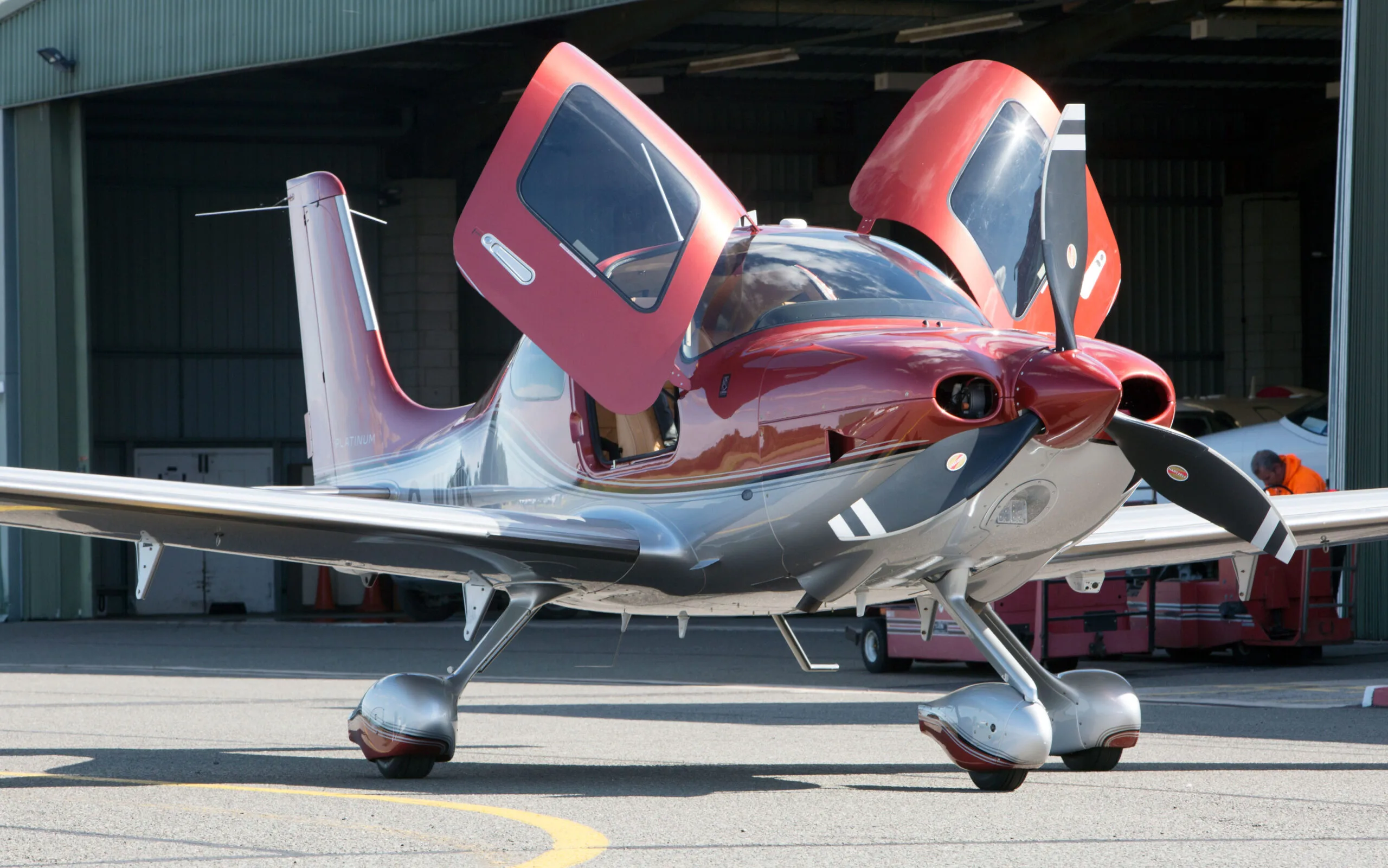 A red and silver Cirrus SR22T GTS aircraft with its gull-wing doors open, parked on an airfield tarmac in front of a hangar.
